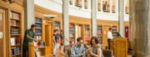 The University of Leeds. Brotherton library. Staff member placing books onto shelves. Library member checking a book out. Two women walking through the library in conversation. Man and women looking at a tablet. Women in background on balcony.
